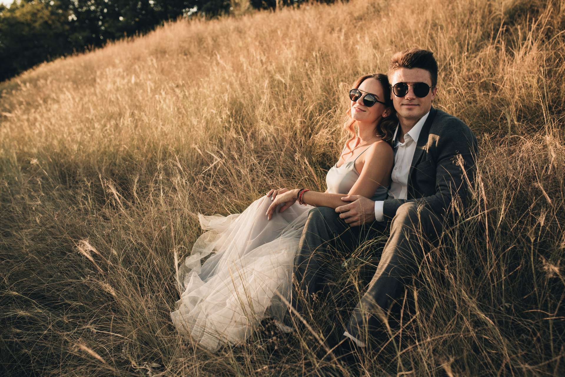 A groom and bride wearing glasses sit in a field surrounded by tall grass. Captured by wedding photographer Vancouver